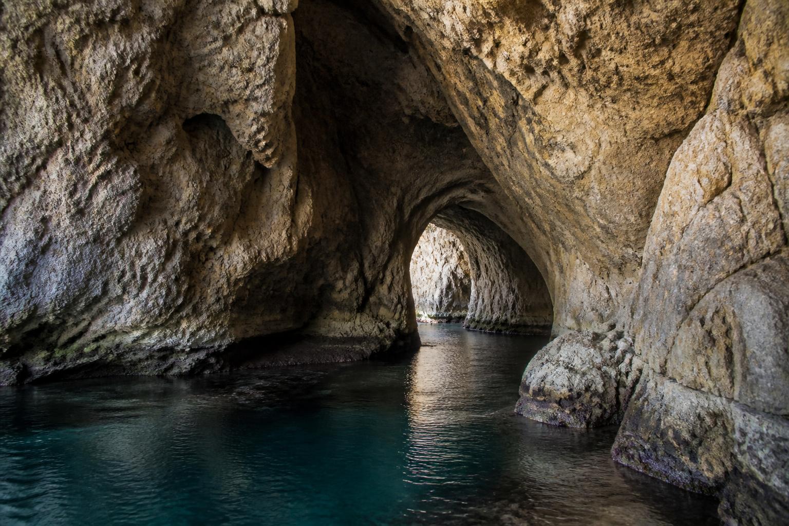 Sunlit coastal cave interior with calm water and rugged stone walls.